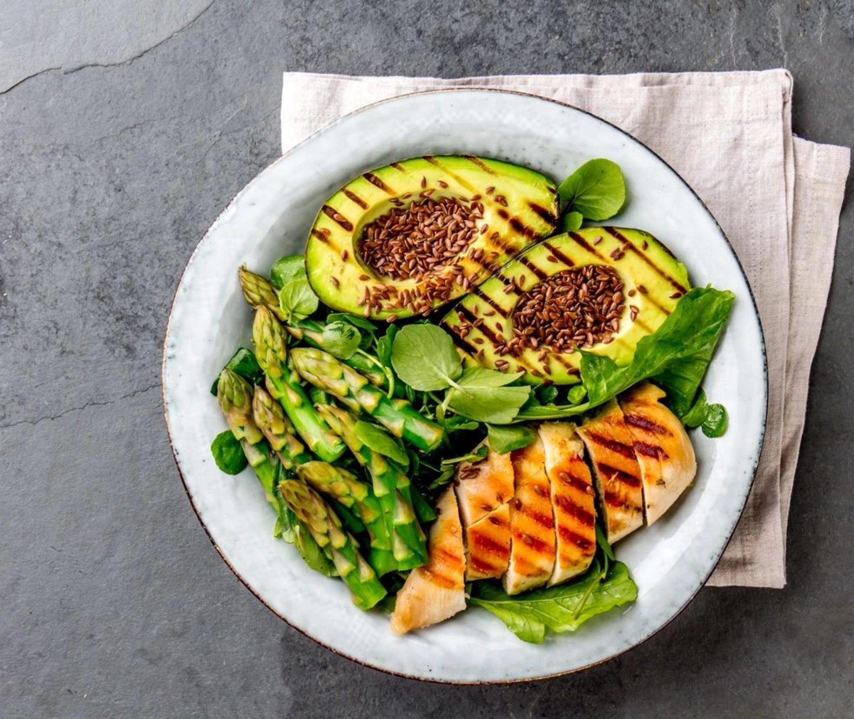 A white bowl of food with grilled chicken, asparagus and peas.